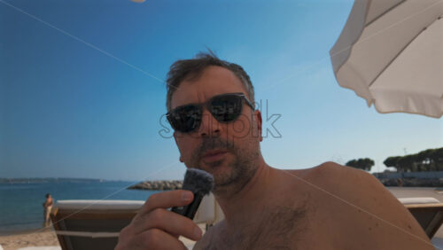 Video - Man wearing sunglasses speaks into a small microphone under large white beach umbrellas with palm trees in the background