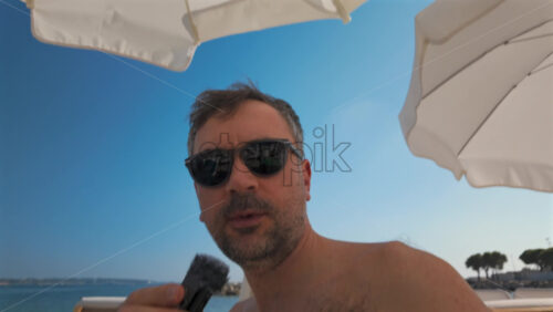 Video - Man wearing sunglasses speaks into a small microphone under large white beach umbrellas with palm trees in the background