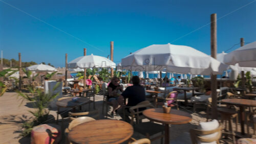 Video - Cannes, France - October 3, 2025: Beachfront cafe with wooden tables, chairs, and white umbrellas under a bright blue sky