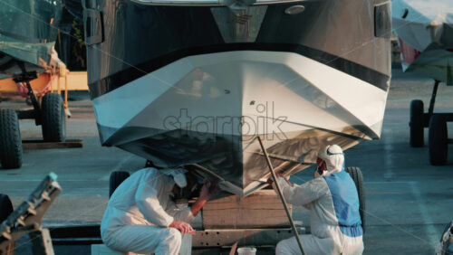 Video - Cannes, France - October 11, 2025: Two workers wearing protective white suits and respirators sand and repair the hull of a boat in dry dock