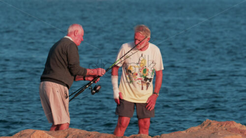 Video - Cannes, France - October 11, 2025: Two elderly men stand on a rocky shore fishing together at sunset, surrounded by calm blue water