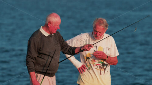 Video - Cannes, France - October 11, 2025: Two elderly men stand on a rocky shore fishing together at sunset, surrounded by calm blue water