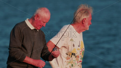 Video - Cannes, France - October 11, 2025: Two elderly men stand on a rocky shore fishing together at sunset, surrounded by calm blue water