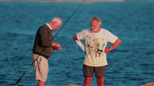 Video - Cannes, France - October 11, 2025: Two elderly men stand on a rocky shore fishing together at sunset, surrounded by calm blue water