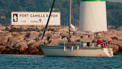 Video - Cannes, France - October 11, 2025: A sailboat with several people onboard approaches a rocky pier beside a green and white lighthouse