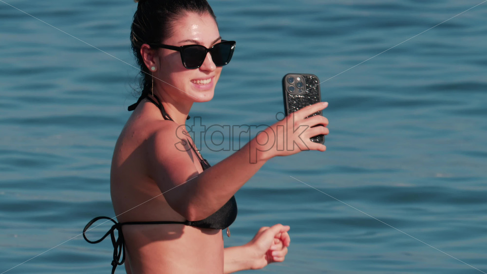 Video - A young woman wearing sunglasses and a black bikini smiles while taking a selfie in the water