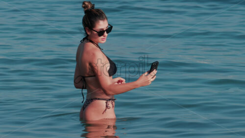 Video - Cannes, France - October 11, 2025: A young woman wearing sunglasses and a black bikini smiles while taking a selfie in the water