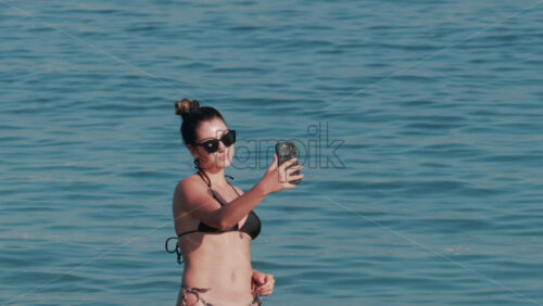 Video - Cannes, France - October 11, 2025: A young woman wearing sunglasses and a black bikini smiles while taking a selfie in the water