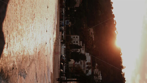 Video - The sun sets over a coastal town with trees, hills, and buildings reflected on the golden sea. Vertical