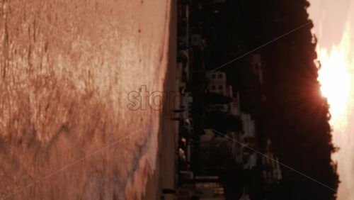 Video - The sun sets over a coastal town with trees, hills, and buildings reflected on the golden sea. Vertical