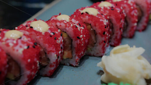 Video - Close up of vibrant red sushi rolls topped with creamy sauce and sesame seeds, served on a ceramic plate
