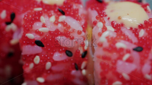 Video - Close up of vibrant red sushi rolls topped with creamy sauce and sesame seeds, served on a ceramic plate