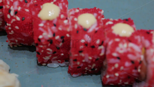 Video - Close up of vibrant red sushi rolls topped with creamy sauce and sesame seeds, served on a ceramic plate