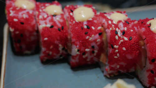 Video - Close up of vibrant red sushi rolls topped with creamy sauce and sesame seeds, served on a ceramic plate