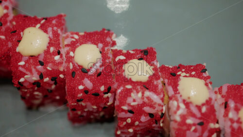 Video - Close up of vibrant red sushi rolls topped with creamy sauce and sesame seeds, served on a ceramic plate