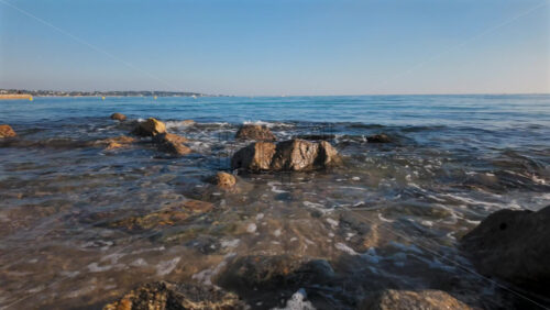 Video - Small waves gently breaking on rocks near the beach, with a view of the coastline and distant buildings