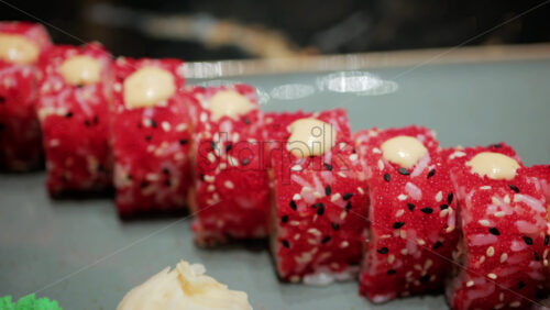 Video - Close up of vibrant red sushi rolls topped with creamy sauce and sesame seeds, served on a ceramic plate