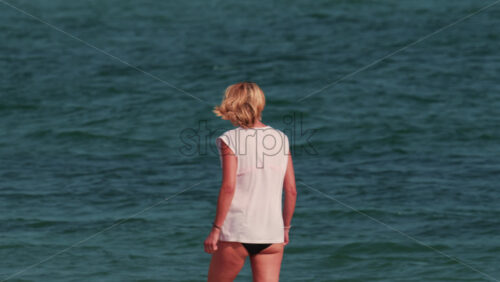 Video - A woman with short blond hair stands waist deep in the sea, facing the horizon