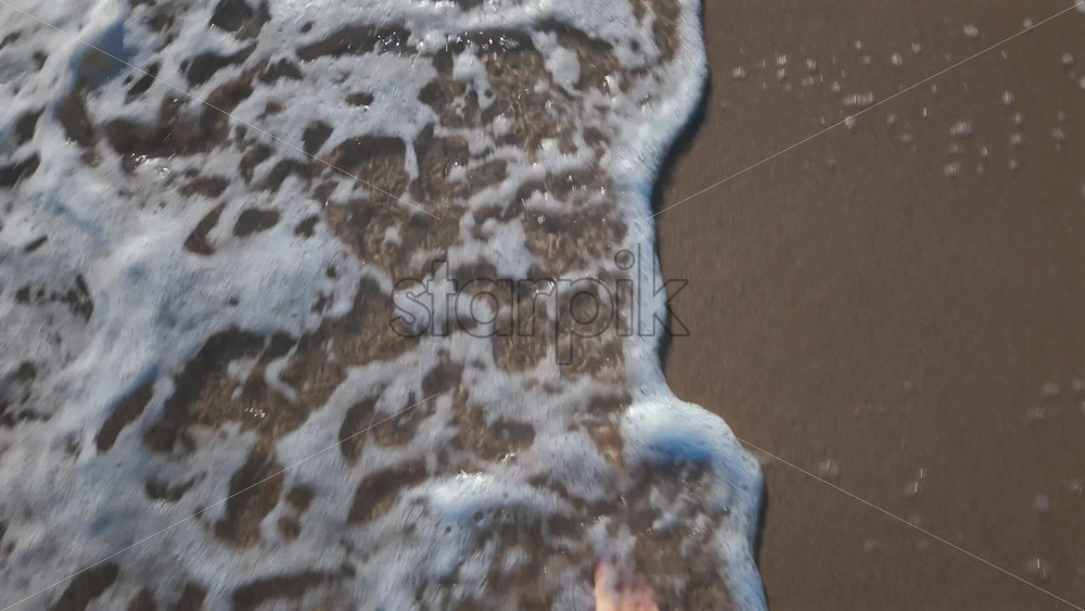 Video - Close up of a man's bare foot touching sea foam while walking on the sandy beach