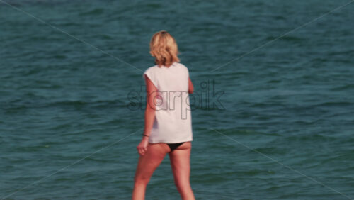Video - A woman with short blond hair stands waist deep in the sea, facing the horizon