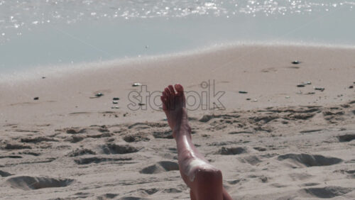 Video - Close up of a woman's legs stretched on the beach as gentle waves roll in