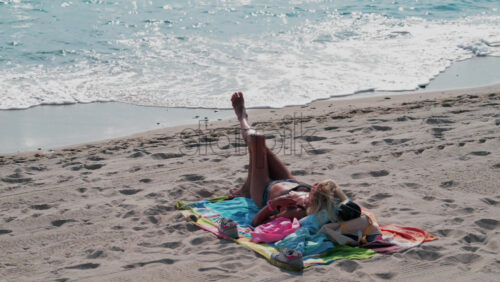 Video - A woman in a bikini relaxes on a colorful towel by the sea, sunbathing under bright daylight