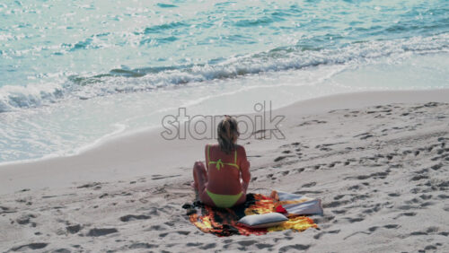 Video - A woman wearing a green bikini sits on a towel facing the ocean, watching the waves sparkle in the sunlight