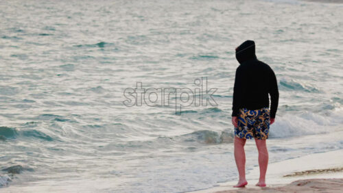 Video - A man wearing a hoodie and patterned shorts stands barefoot by the shoreline, staring at the calm sea