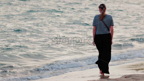 Video - A woman walks barefoot along the beach, waves gently touching her feet
