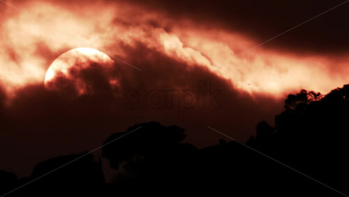 Video - A glowing red sun partially covered by dark clouds at dusk