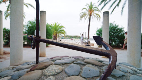Video - A metal anchor displayed on a stone base near palm trees and a seaside promenade