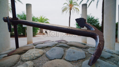 Video - A metal anchor displayed on a stone base near palm trees and a seaside promenade