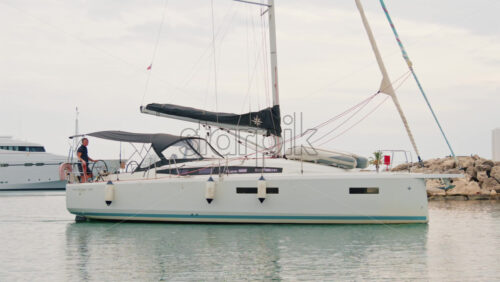 Video - Cannes, France - October 12, 2025: A man steering a white sailboat as it leaves a marina on a calm day
