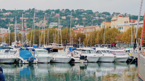 Video - Cannes, France - October 12, 2025: Dozens of yachts and boats docked in a harbor with a colorful Mediterranean town and green hills in the background