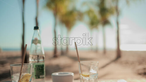 Video - Two refreshing drinks with lemon slices and straws on a table overlooking a sunny beach with palm trees