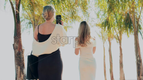 Video - Two women in elegant summer outfits taking photos under palm trees near the beach