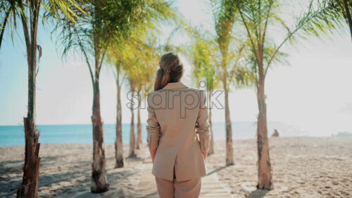 Video - Stylish woman wearing a beige suit and sunglasses walks calmly along a palm lined wooden path toward the sea