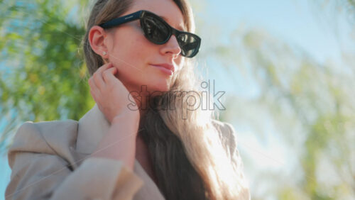 Video - Portrait of a woman in sunglasses and beige suit looking up toward the sky with palm trees in the background
