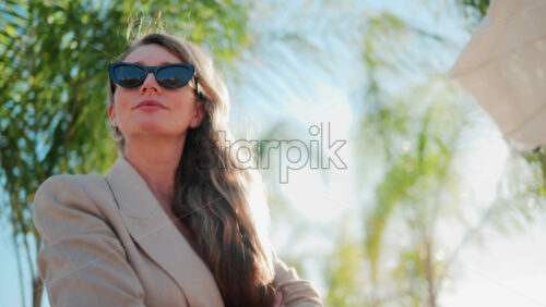 Video - Stylish woman wearing a beige suit and sunglasses walks calmly along a palm lined wooden path toward the sea