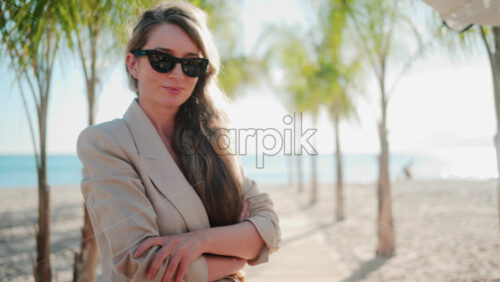Video - Stylish woman wearing a beige suit and sunglasses walks calmly along a palm lined wooden path toward the sea