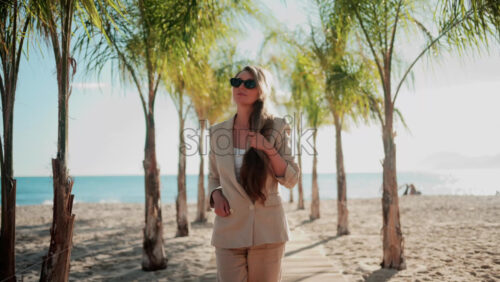 Video - Stylish woman wearing a beige suit and sunglasses walks calmly along a palm lined wooden path toward the sea