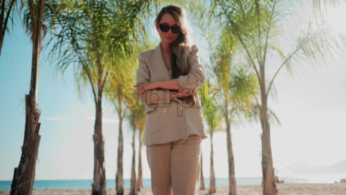 Video - Stylish woman wearing a beige suit and sunglasses walks calmly along a palm lined wooden path toward the sea