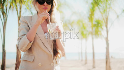 Video - Elegant woman holding a glass of lemon water while relaxing at a beach bar under palm trees
