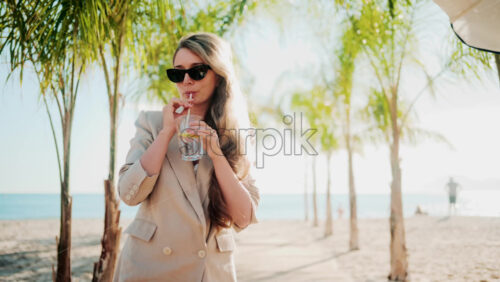 Video - Elegant woman holding a glass of lemon water while relaxing at a beach bar under palm trees