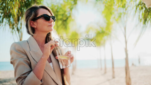 Video - Elegant woman holding a glass of lemon water while relaxing at a beach bar under palm trees