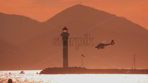 Video - Helicopter flying past a lighthouse with orange sunset sky and distant mountains