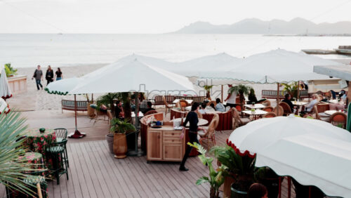 Video - Cannes, France - October 13, 2025: People enjoying drinks and conversation at a stylish beach bar with large umbrellas and tropical plants