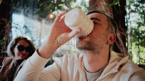 Video - Close up of a man sipping coffee in natural sunlight, near a woman wearing sunglasses at a cozy outdoor cafe
