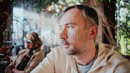 Video - Close up of a man sipping coffee in natural sunlight, near a woman wearing sunglasses at a cozy outdoor cafe