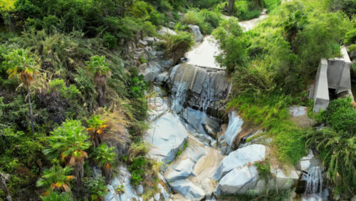 Video – Aerial drone view of a small waterfall cascading over smooth rocks, surrounded by palm trees and wild plants in slow motion - Starpik Stock